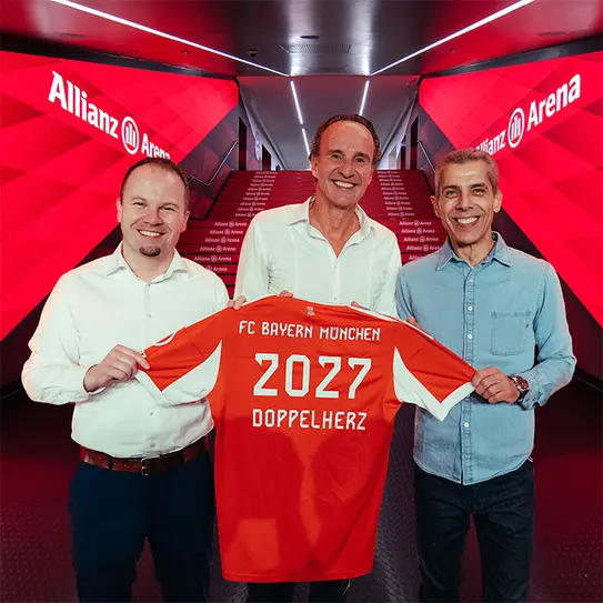 三位男士在安联竞技场前合影,展示一件印有“FC Bayern München 2027 DOPPELHERZ”的足球衣。背景为红色灯光和竞技场标志,传达出兴奋和庆祝的氛围。